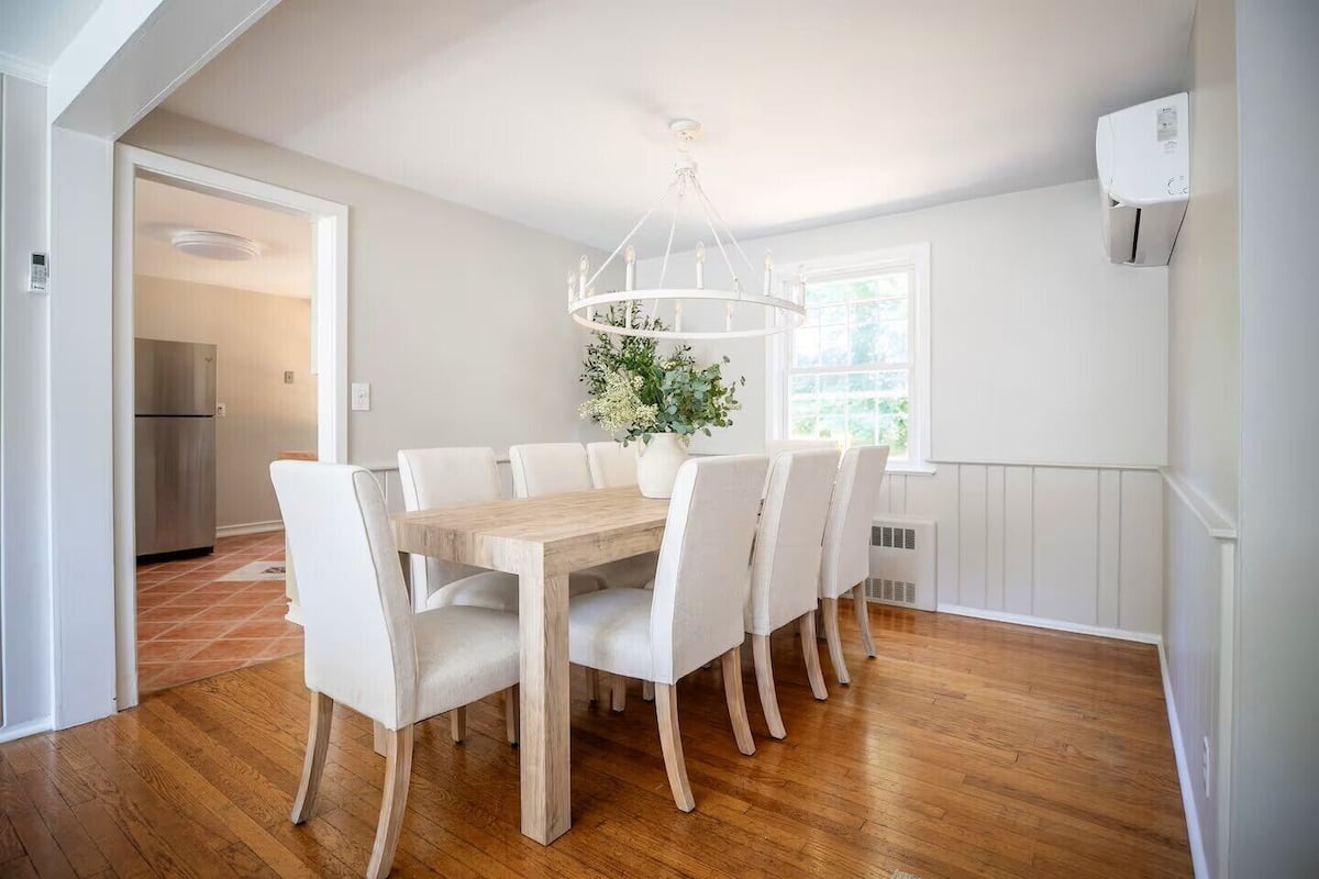 Un coin repas avec une table en bois et des chaises blanches, un lustre et une vue sur la cuisine.