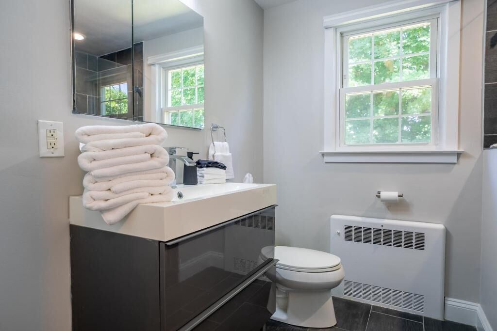 A modern bathroom with a white sink, a toilet, a towel rack with folded towels, a mirror, and a window with a view of greenery.