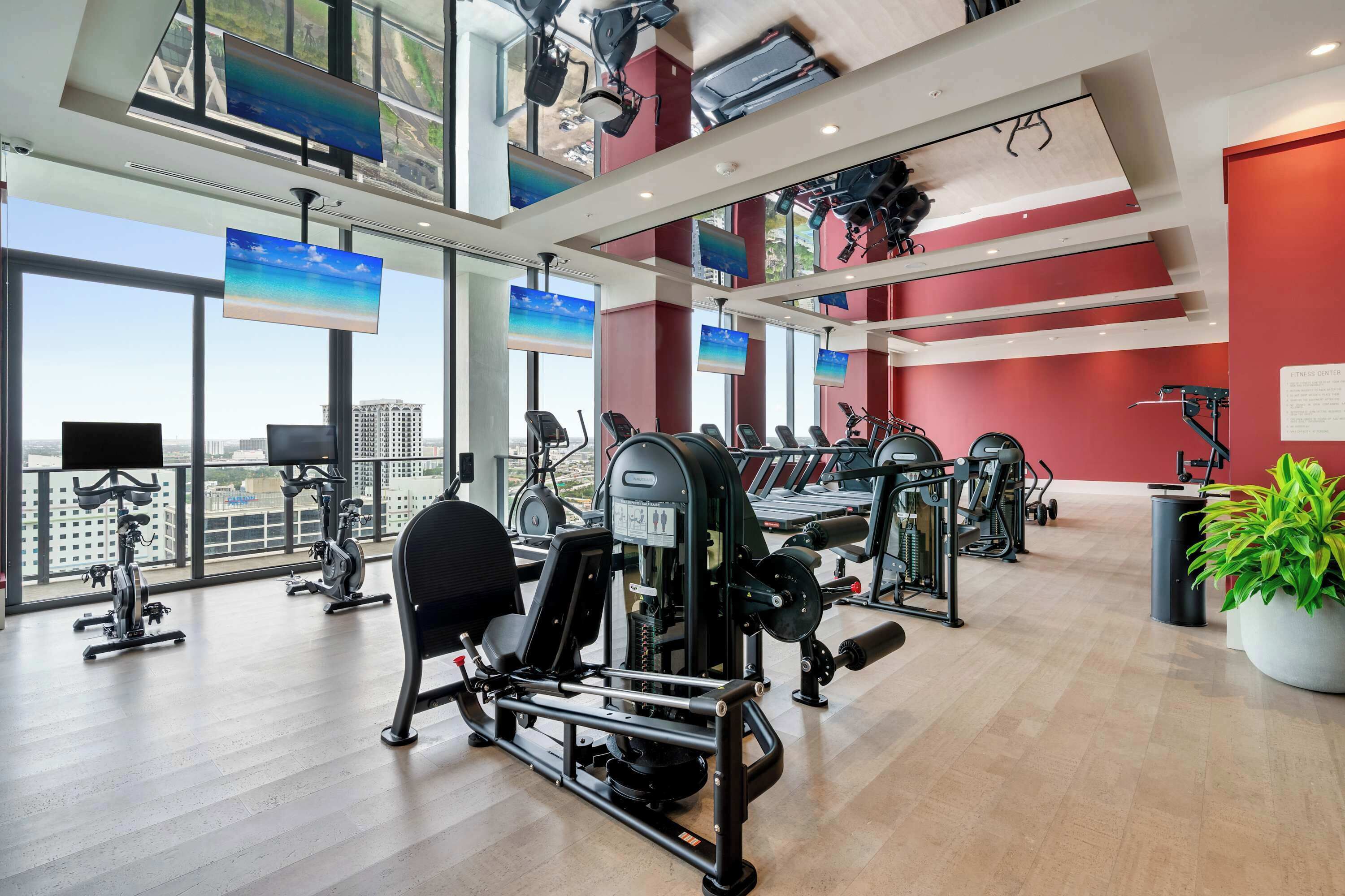 Une salle de sport équipée de divers appareils d’exercice, dotée de grandes fenêtres et offrant une vue sur la ville.