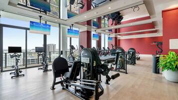 Une salle de sport équipée de divers appareils d’exercice, dotée de grandes fenêtres et offrant une vue sur la ville.