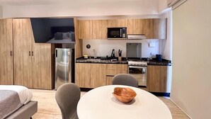 A compact kitchen with wooden cabinets, a refrigerator, microwave, and a stove. There is a round table with a bowl and two chairs. - Mi Zona Lodging Chapultepec (Mexico City)