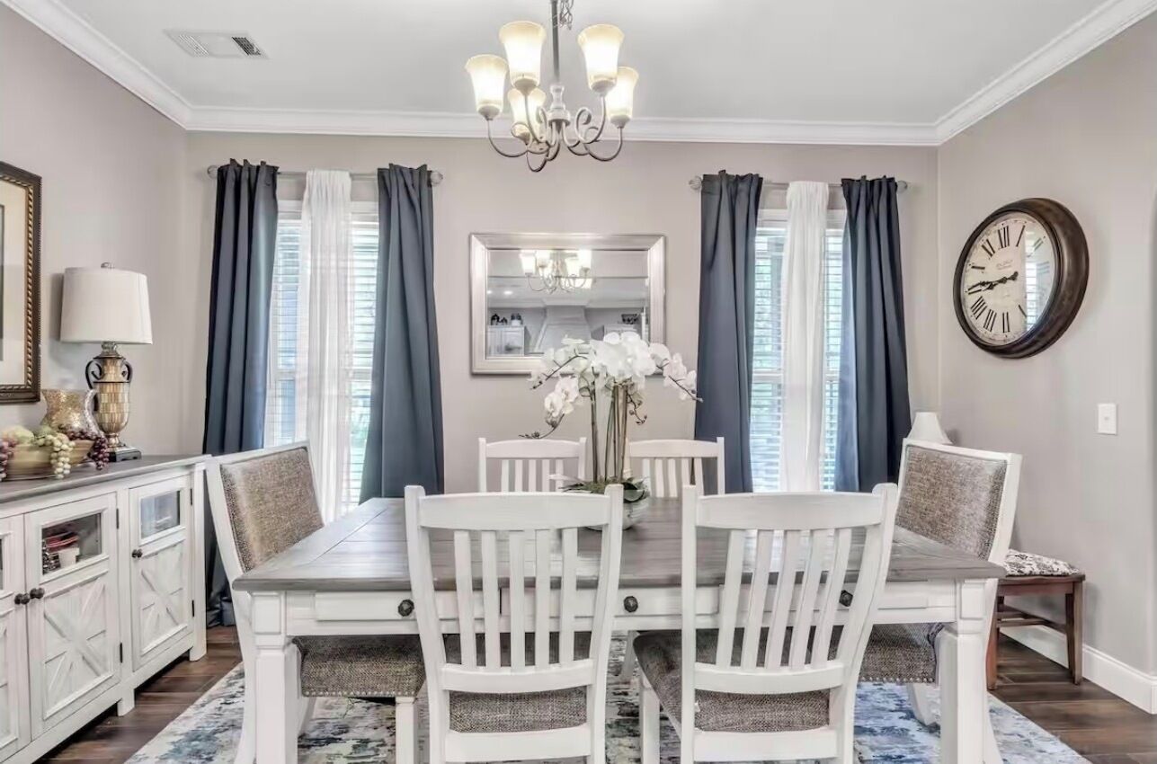 Une salle à manger avec une table blanche, quatre chaises, un lustre et une horloge au mur.