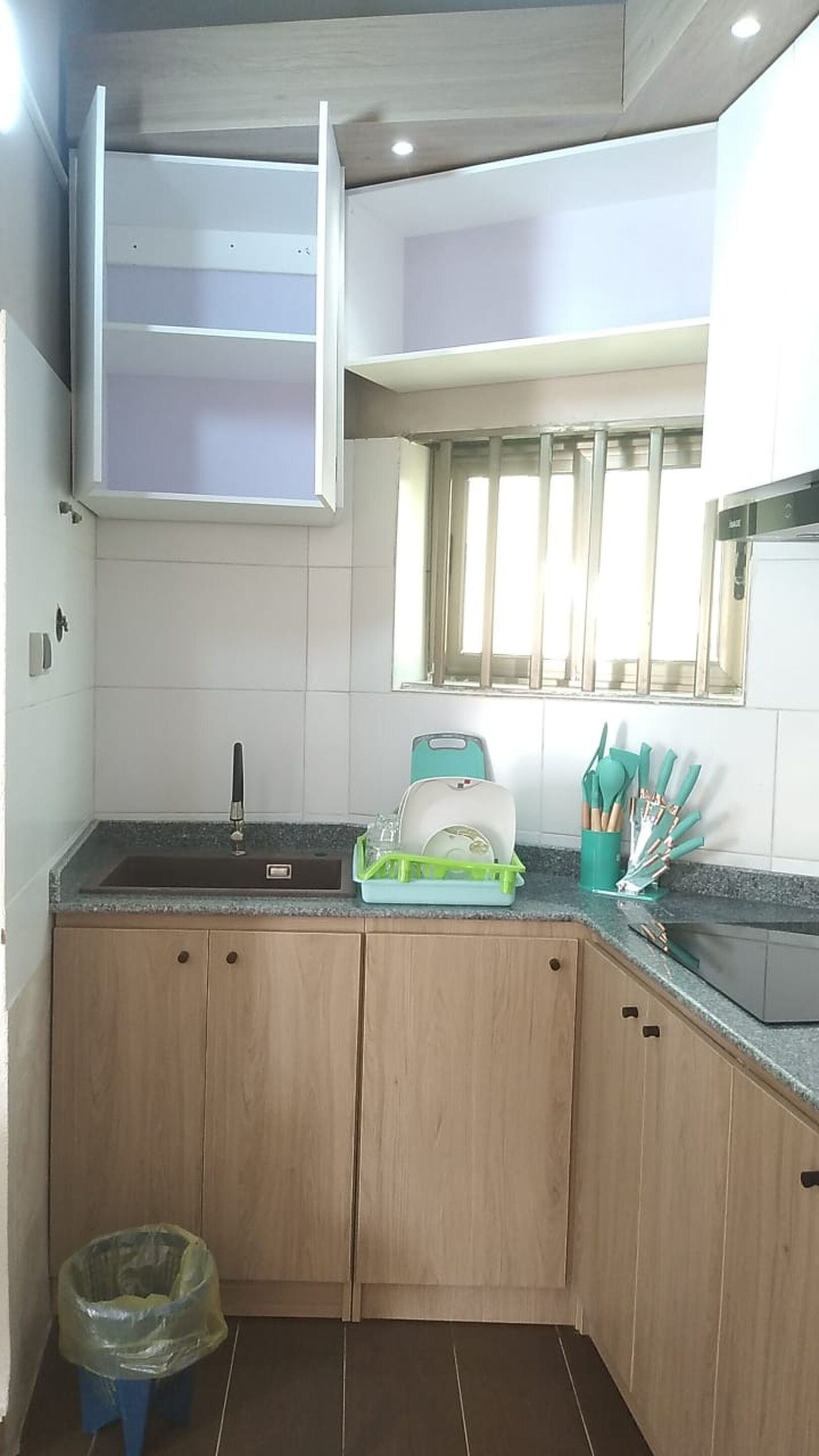 Cocina compacta con lavavajillas integrado, una ventana con persianas y un pequeño cubo de basura.