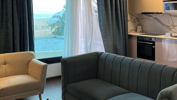 Una sala de estar moderna con un sofá, sillones y una mesita de centro. Cuenta con una zona de cocina con microondas y estufa. La habitación tiene ventanas amplias con cortinas, y ofrece vistas a la playa.