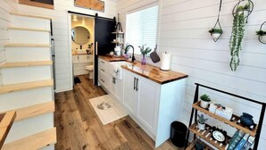 A modern kitchen with white cabinetry, a wooden countertop, and a black backsplash. There are hanging plants, a small shelf with kitchen items, and a window with blinds. - Private Hot Tub + Rooftop Deck • Near Zion National Park (Apple Valley)