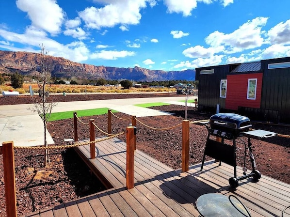 Une terrasse en bois avec un barbecue, un petit bâtiment rouge et, au loin, une vue sur la montagne.