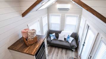 Een knus interieur met een houten plafond, een grijze bank met gestreepte kussens, een mand met spullen en een raam met zonwering.