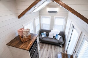 A cozy room with a wooden ceiling, a gray sofa with patterned cushions, a basket with items, and a window with blinds. - Tiny Home + Private Hot Tub | Zion Adventure Base (Apple Valley)