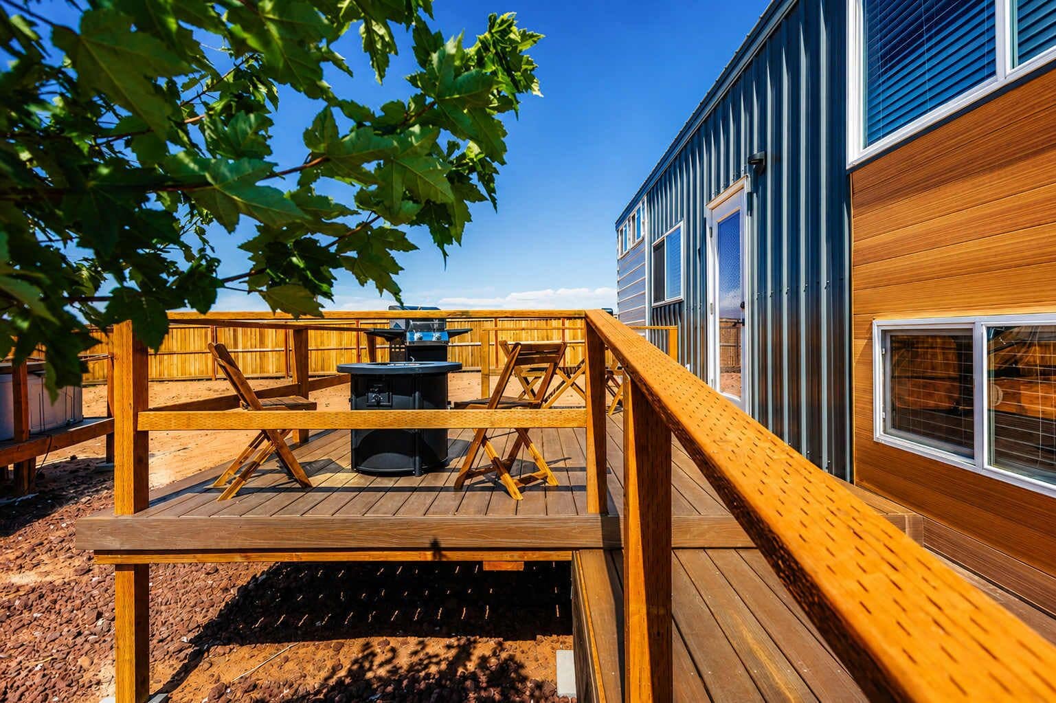 Eine Holzterrasse mit Grill und Stühlen, ein Holzzaun und ein Gebäude mit großen Fenstern.