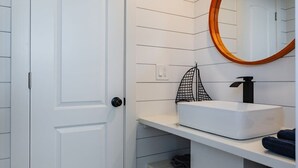 A bathroom with a white door, a round mirror with a wooden frame, a white sink, and a shelf with towels. - Zion Tiny Home Retreat w/ Hot Tub & Deck (Apple Valley)