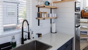 A modern kitchen with a large island, stainless steel sink, and built-in appliances. There are wooden shelves above the island and a window with blinds. - Zion Tiny Home Retreat w/ Hot Tub & Deck (Apple Valley)