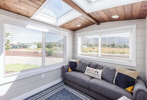 Ein Zimmer mit grauem Sofa, zwei Fenstern mit Jalousien, Holzboden und Blick auf eine Wohngegend.