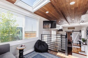 Ein modernes Interieur mit Holzboden, einem großen Fenster mit Jalousien, einem schwarzen Sitzsack, einem kleinen Tisch mit Zeitschriften und einem an der Wand befestigten Fernseher.