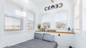 Un dormitorio con cama, dos ventanas, un reloj de pared, una planta pequeña y un ventilador de techo.
