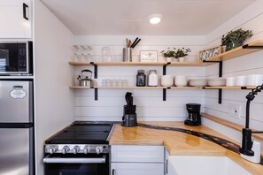 Une cuisine moderne équipée d’une cuisinière, d’un four et d’un four à micro-ondes intégrés. On y trouve des étagères en bois où sont rangés les ustensiles de cuisine, ainsi qu’une machine à café posée sur le plan de travail.