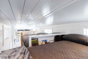 Un dormitorio moderno y compacto con una cama litera y una cama individual, paredes y techo blancos, una colcha acolchada y una ventana pequeña.