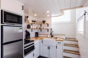 A compact kitchen with a built-in oven, a microwave, and a coffee maker. There are wooden shelves above the counter and a window with blinds. - Zion Basecamp • Hot Tub • Mountain Biking & Hiking (Apple Valley)