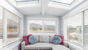 A modern room with a sectional sofa, large windows with blinds, and a skylight. - Hot Tub Under Desert Sky • Zion Tiny Home Getaway (Apple Valley)