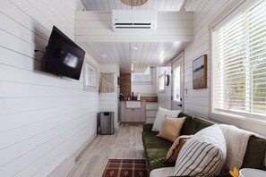 A compact living space with a green sofa, a flat-screen TV mounted on the wall, a kitchen area, and a window with blinds. - Pet-Friendly Zion Group Getaway (Apple Valley)