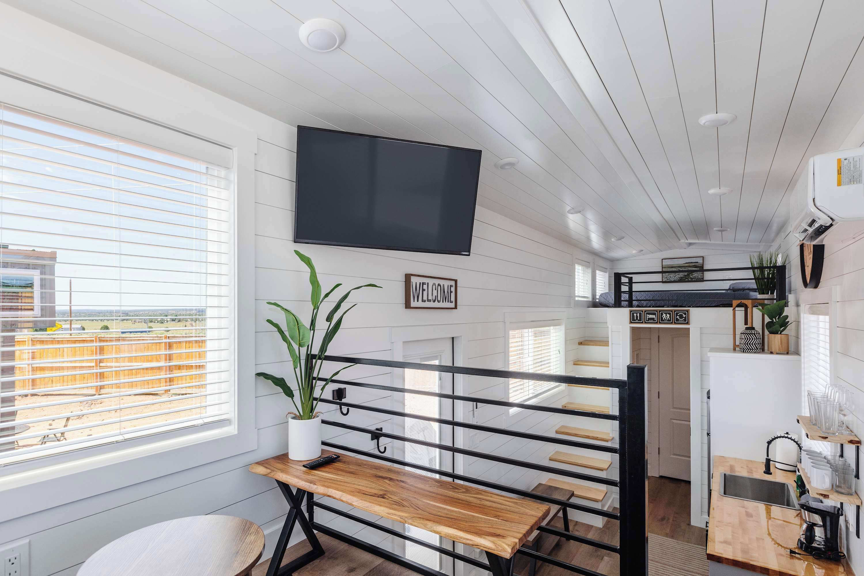 Ein modernes Interieur mit an der Wand montiertem Fernseher, einer Bank, einem Blumentopf und einer Küchenzeile mit Spüle und Arbeitsplatte.