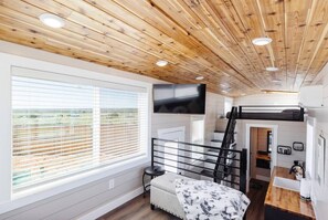 Een moderne, compacte kamer met een stapelbed, een groot raam met zonwering, een flatscreen televisie en een klein keukenhoekje.