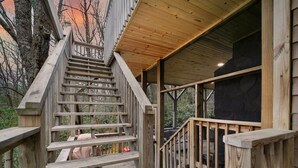 Eine Holztreppe führt zu einer überdachten Terrasse mit Blick auf Bäume und einen Sonnenuntergang.