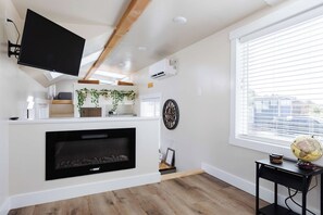 Un salon moderne avec une cheminée, un téléviseur fixé au mur et une grande fenêtre équipée de stores.