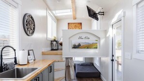 Eine kleine Küche mit Spüle, Schränken und einer Holzauszugplatte. Es gibt eine kleine Sitzecke mit einer Bank und einem an der Wand befestigten Fernseher.
