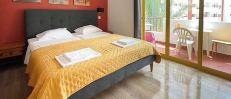 A hotel room with a bed, a balcony with chairs, and a view of buildings.