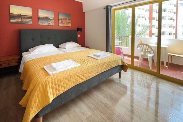 A hotel room with a bed, a balcony with chairs, and a view of buildings.