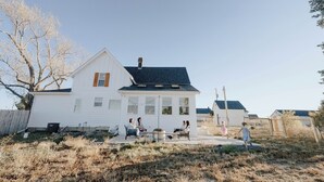 A white house with a dark roof and a chimney, surrounded by dry grass and a wooden fence.