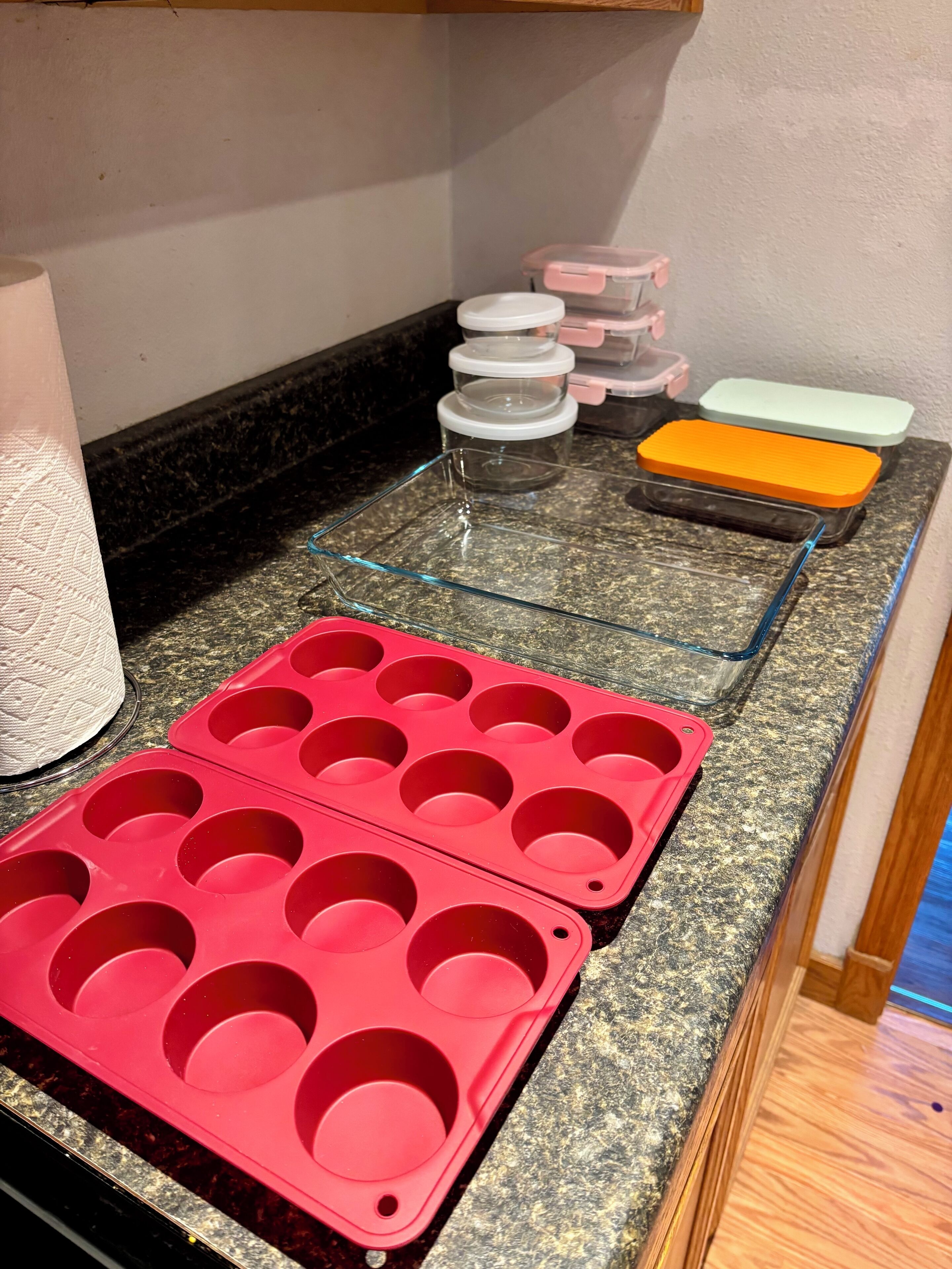 A kitchen counter with a red muffin tray, a glass baking dish, and a stack of clear plastic containers.