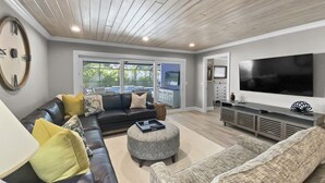 A modern living room with a black leather sofa, patterned ottoman, and grey couch. A flat-screen TV is mounted on the wall, and there is a large window providing a view of the outdoors. - Modern Luxury Private Home with Pool in Siesta Key (Siesta Key)