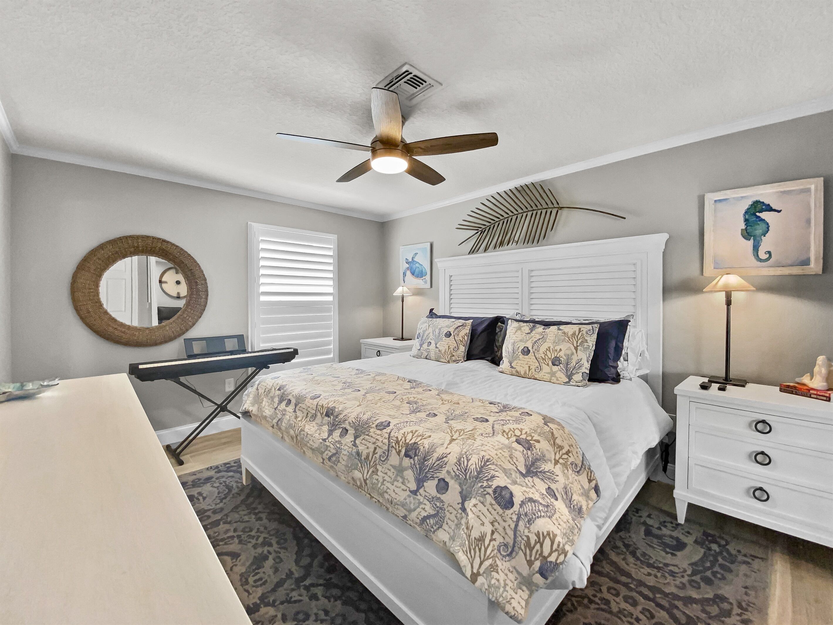 Une chambre à coucher comprenant un lit, un ventilateur de plafond, un bureau, une chaise, un miroir, une fenêtre avec des stores et une table de chevet avec une lampe.
