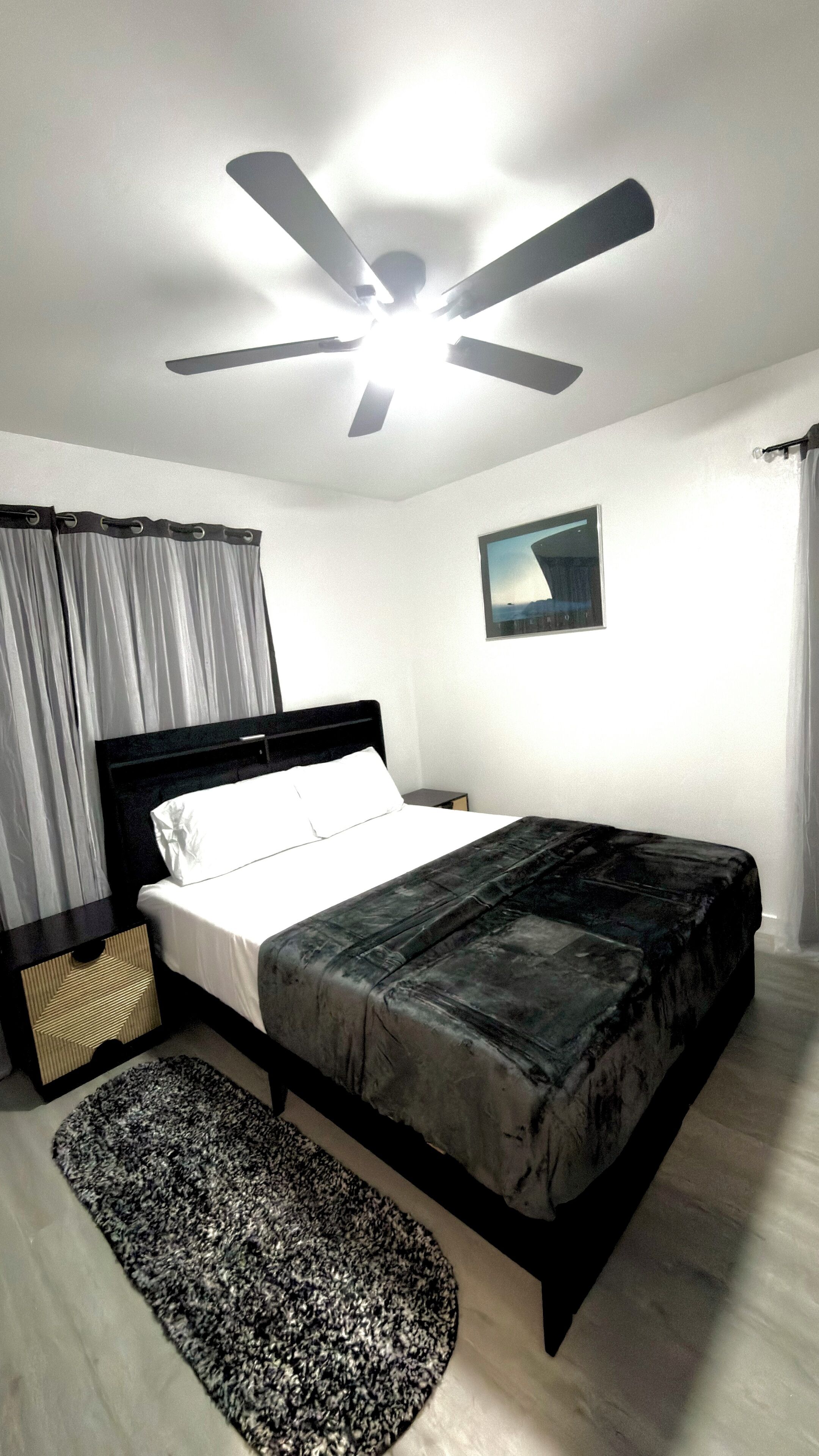 Quarto com cama, ventoinha no teto, mesa de cabeceira, tapete e um quadro na parede.