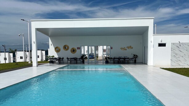 A modern pool area with a clear blue pool, white walls, and a covered seating area with tables and chairs.