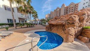 A swimming pool with a rock formation, palm trees, and a building in the background. - Delphin 103, Las Palmas Resort (Puerto Peñasco)