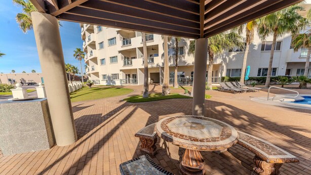 A covered outdoor area with a circular table and benches, palm trees, and a building with balconies in the background. - Delphin 103, Las Palmas Resort (Puerto Peñasco)