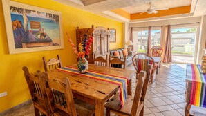 A dining area with a wooden table, chairs, a colorful table runner, a painting of a beach scene, and a view through the glass doors. - Delphin 103, Las Palmas Resort (Puerto Peñasco)