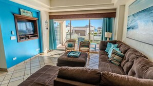 A living room with a brown sofa, a television, and a view of a house and ocean. - Grande 105, Las Palmas Resort (Puerto Peñasco)