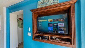 A living room with a TV mounted on a blue wall, a wooden shelf below, and a sign above the TV that reads 'BEACH This is where I belong.' - Grande 105, Las Palmas Resort (Puerto Peñasco)