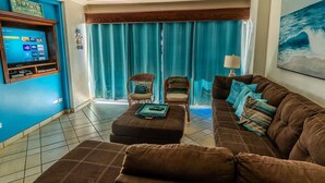 Un salon avec un grand canapé marron, une table basse et deux fauteuils. Un téléviseur à écran plat est fixé au mur, et il y a une fenêtre avec des rideaux bleus.