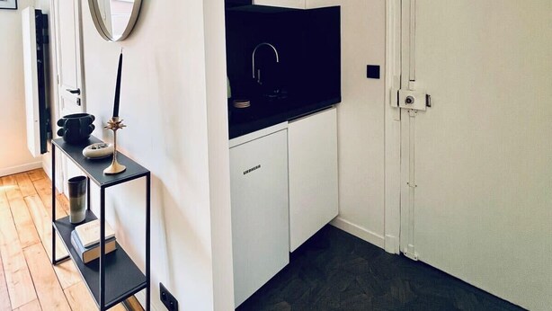 A modern kitchen with white cabinets, a black countertop, and a built-in stove. There is a black metal shelf with a vase and a book. A round mirror hangs on the wall above the shelf. - Bastille · 11th · Stylish & Quiet Studio – managed by Mey Agency (Paris)