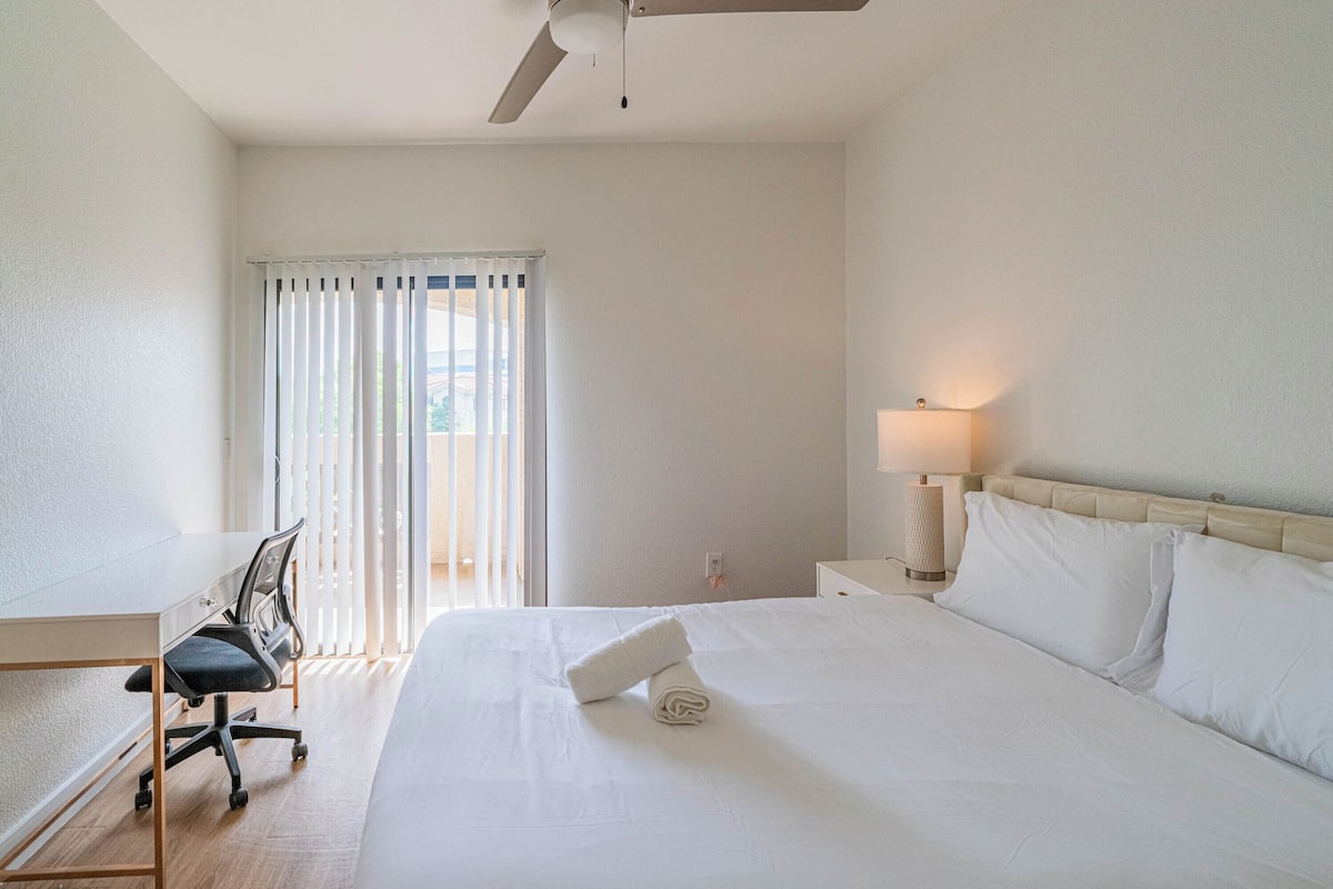 Una camera d'albergo con un letto, una scrivania, una sedia, una lampada e una finestra con tende.