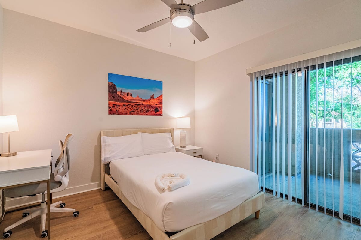 Una camera d'albergo con un letto, una scrivania, una sedia, una ventola a soffitto, un comodino, una lampada, un quadro e un'ampia finestra con tende.