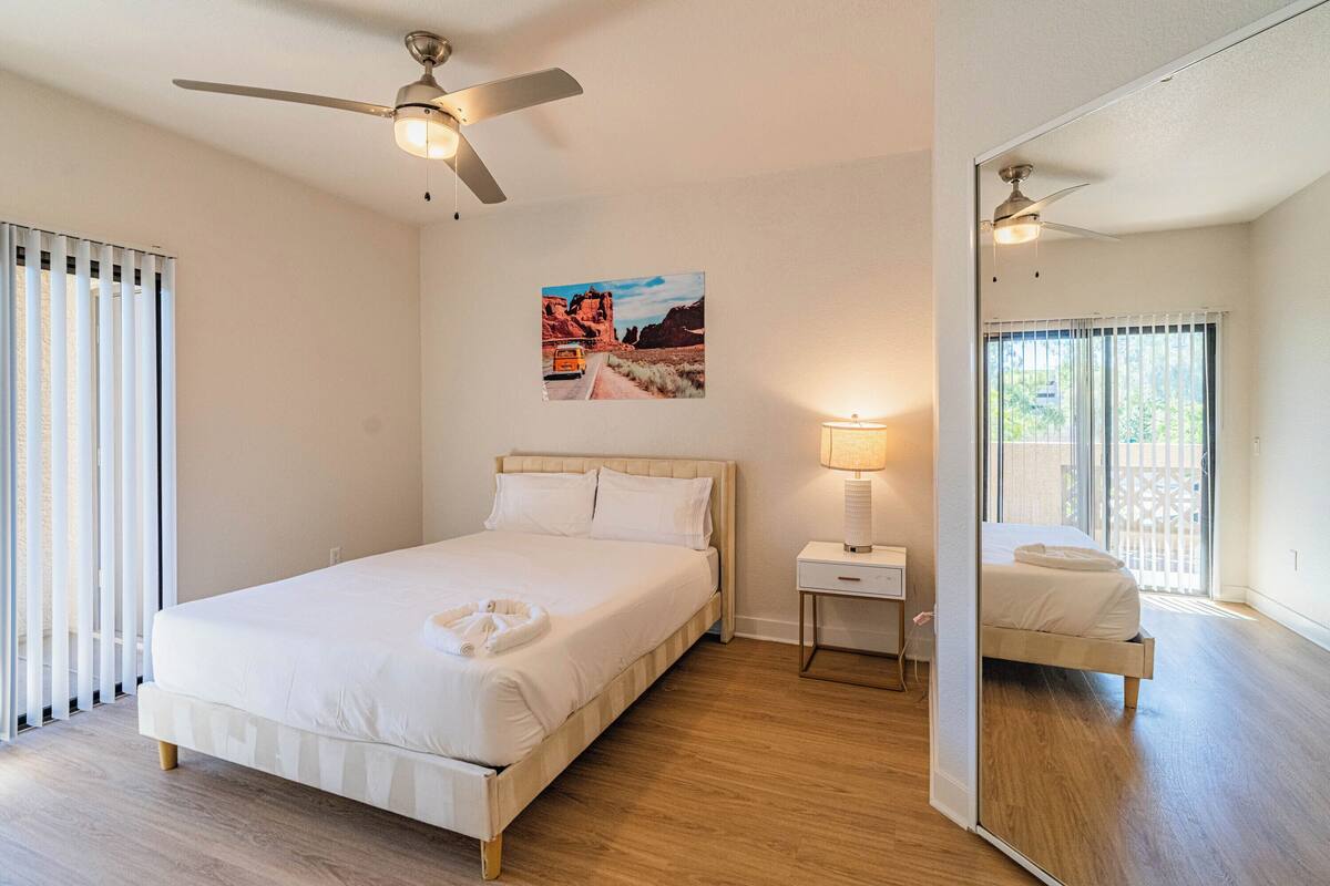 Una camera da letto con un letto, una ventola a soffitto, un comodino con una lampada e un'ampia finestra con persiane.