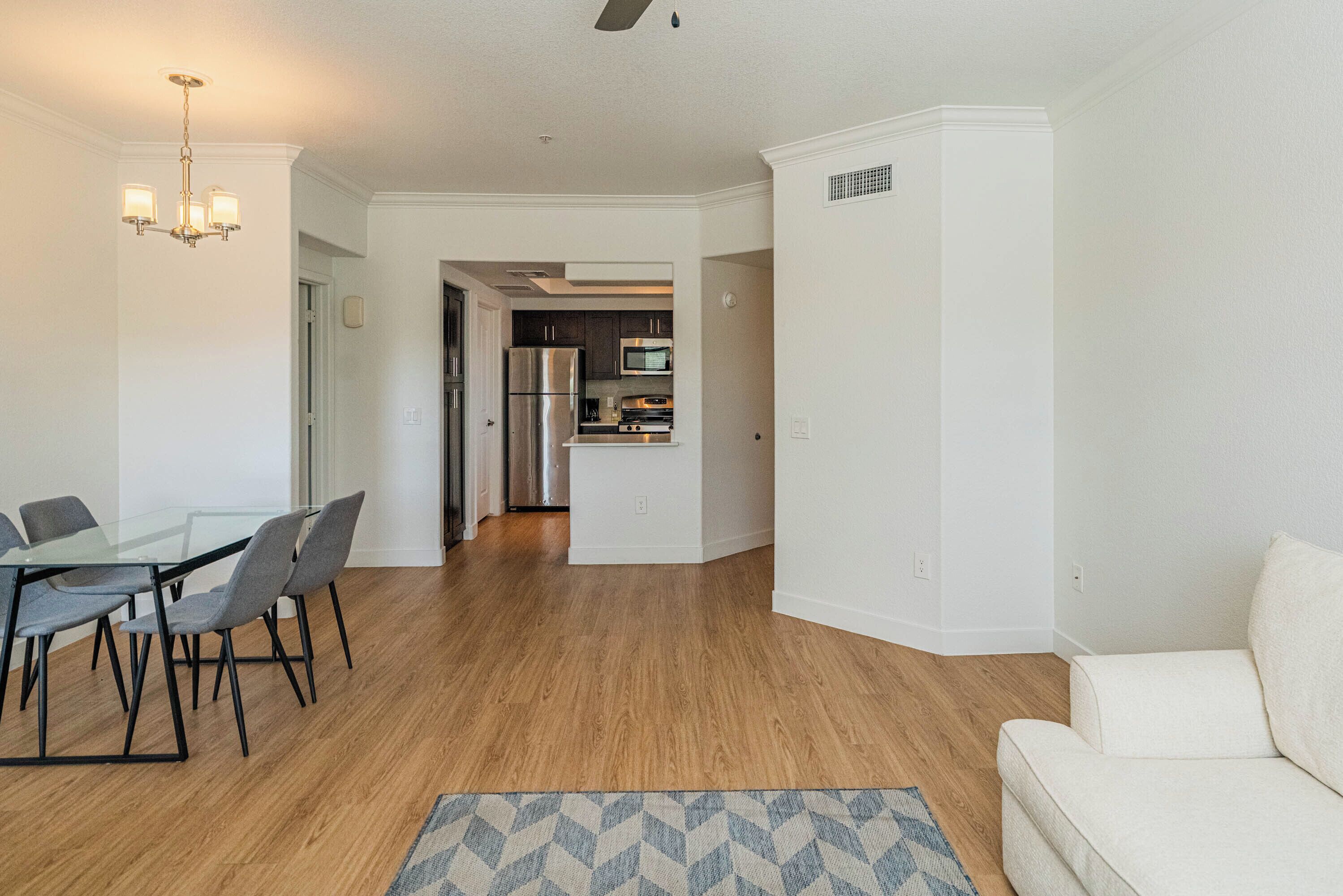 A modern living room with a glass dining table, grey chairs, a white sofa, and a kitchen in the background.