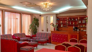 Eine Hotellobby mit roten Sofas, einem Essbereich mit roten Tischen, einer Chandelier und einer Bar mit Regalen voller Flaschen und gerahmten Bildern.