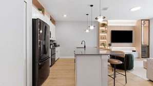 A modern kitchen with a large refrigerator, a central island, pendant lights, and a flat-screen TV mounted on the wall.