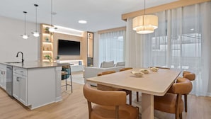 A modern kitchen with a central island, a dining area with a wooden table and chairs, and a living space with a sofa and TV.
